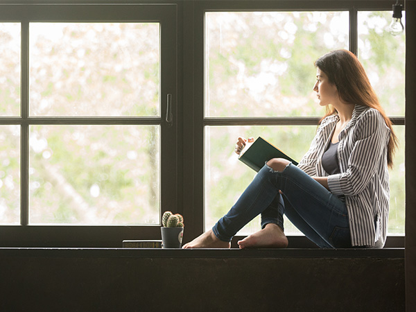 A woman pausing from reading her book to gaze out the window while practicing what she has learned from Acceptance and Commitment Therapy (ACT).