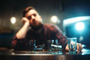 "Alcohol use." Focus on one of many empty shot glasses in front of an out of focus man slumped in a bar.