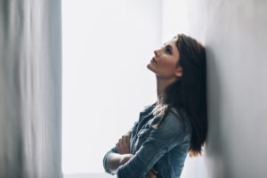 "Anxiety". A stressed young woman leans against a wall, arms crossed, and looks upward as though to calm down.