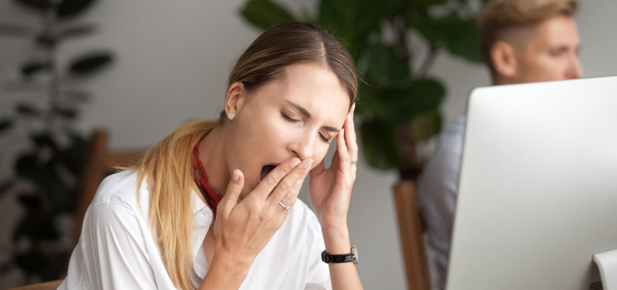 "Can CBT Relieve Symptoms Of Insomnia?". A woman yawning in front of her computer at the office.