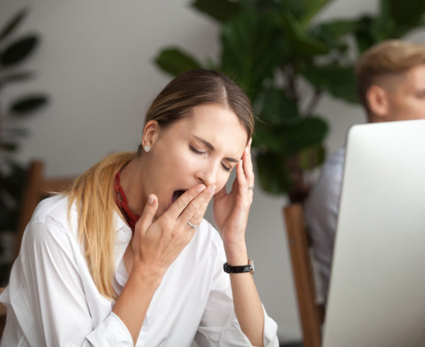 "Can CBT Relieve Symptoms Of Insomnia?". A woman yawning in front of her computer at the office.