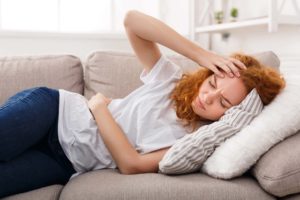 "Chronic health conditions". A woman lays on the couch, holding her head and her sides.