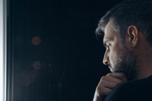 "Depression." A middle aged man strokes his beard in a dark room while gazing down out of a window.