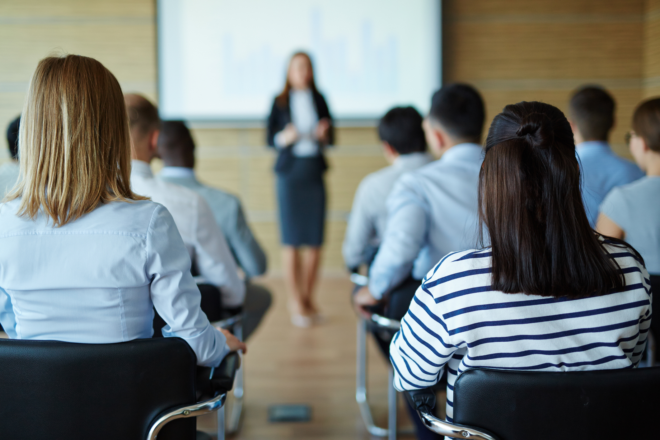 "Public speaking". An interested audience listens to a woman out of focus at the front of a seminar.