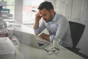 "Stress" - a man in the office with his hand on furrowed brow, looking at his laptop, discarded eyeglasses next to him.