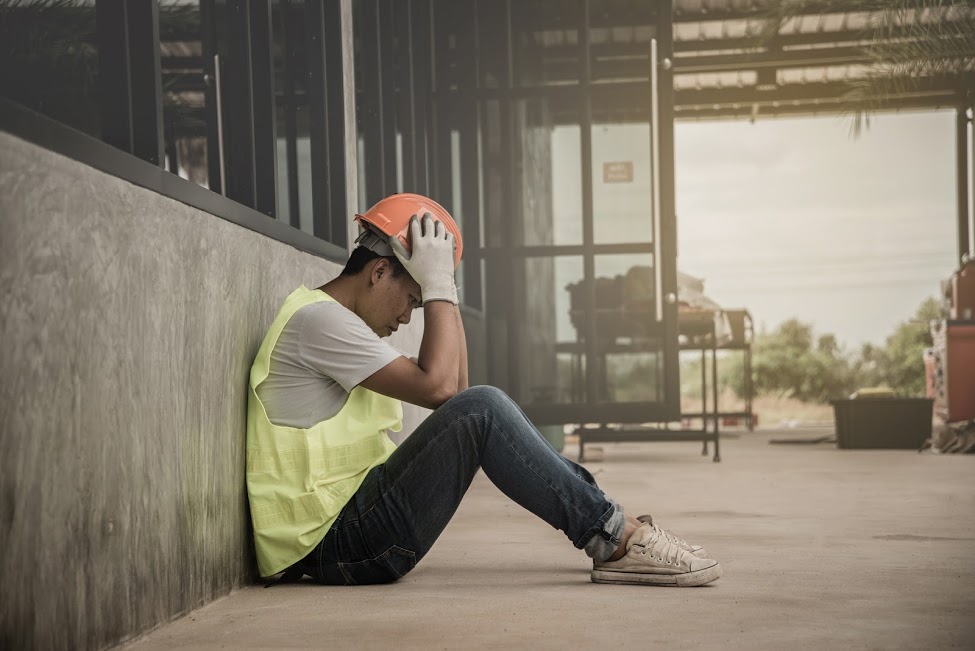 "Agoraphobia". A construction worker holds his helmet while sitting down against a wall, staring at the ground.