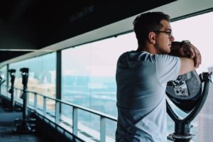 " Grief & Loss Counselling". A man leaning on a viewing device at a skyscraper window while not gazing at the view, wrapped in thought.