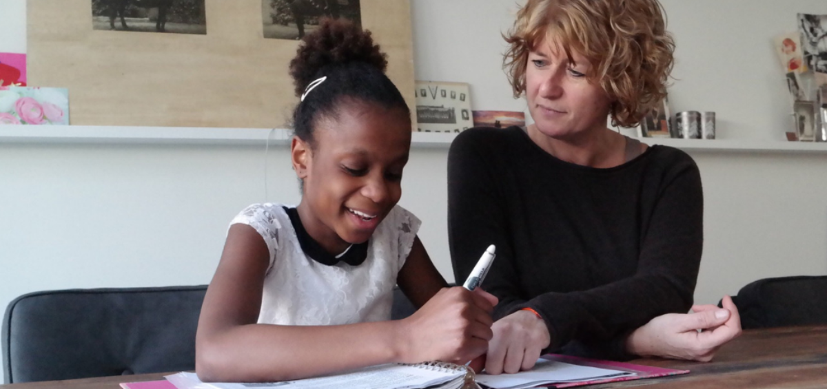 A girl smiles while working in a workbook with a therapist looking on.