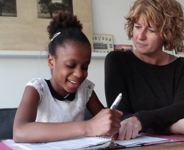A girl smiles while working in a workbook with a therapist looking on.